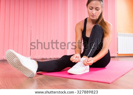 Woman in a gym putting on her trainers as she sits on a pink gym or yoga mat in her sportswear preparing for a workout in a health and fitness concept