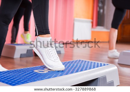 Close Up of Woman Wearing White Sneakers Doing Toe Tap on Step Platform in Aerobic Exercise Class with Group of Women in Background in Dance Studio