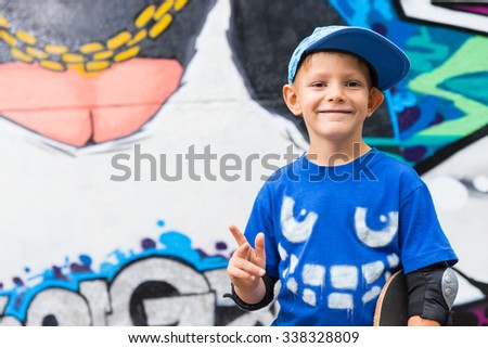 Funny playful kid wearing blue cap and T-shirt with a mean face printed on it posing while pointing up, outdoors in front of a graffiti wall