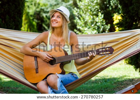 Beautiful blonde woman is sitting on hammock and playing her guitar.Playing guitar