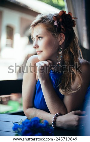 Beautiful young woman sitting at the table. Blonde hair. Blue dress. Flower in hair.