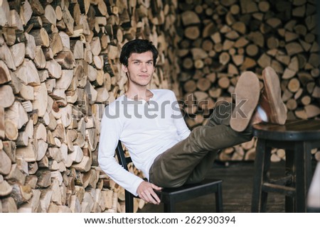 Young man leaning on the back of the chair with his feet up. Firewood background.