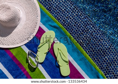 Pair of green flip flops by the pool on a bright blue, green, purple and white striped towel with sunglasses and big white floppy hat