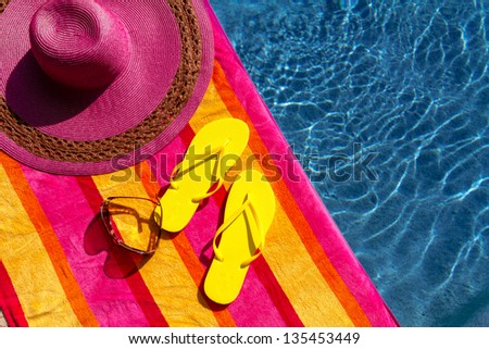 Pair of bright yellow flip flops by the pool on a bright orange, pink, red and yellow striped towel with sunglasses and big pink floppy hat