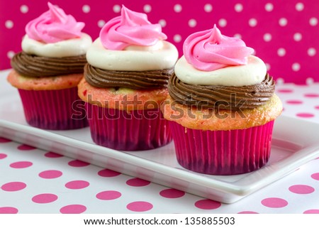 Line up of 3 neapolitan frosted cupcakes on white plate with pink polka dot table and background