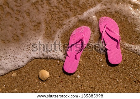 Pair of magenta pink flip flops laying on the sand with ocean wave washing up on the beach