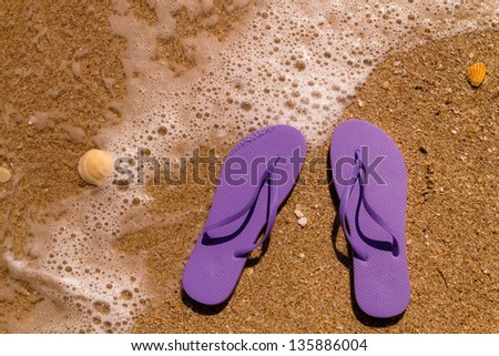 Purple flip flops laying on the sand with ocean water washing up on shore and sea shells