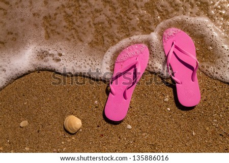 Pink flip flops on a sandy beach with ocean water washing up on shore and sea shells
