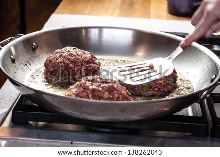 Female chef hand pushing raw hamburger down with spatula in hot stainless steel pan to sear burger
