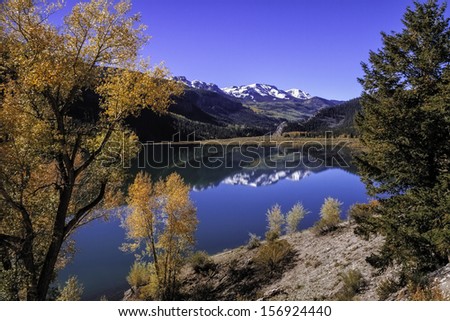 High mountain lake with reflection of snow covered peaks surrounded by changing fall colored trees on autumn afternoon