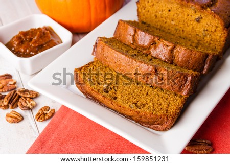 Loaf of pumpkin bread sitting on white plate with orange napkin and container filled with pumpkin butter and pecan nuts