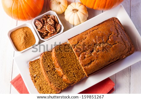 Loaf of pecan pumpkin bread sitting on white plate with orange napkin, spices and assorted pumpkins
