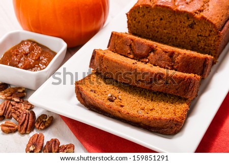 Loaf of pumpkin bread sitting on white plate and container filled with pumpkin butter and pecan nuts with orange napkin