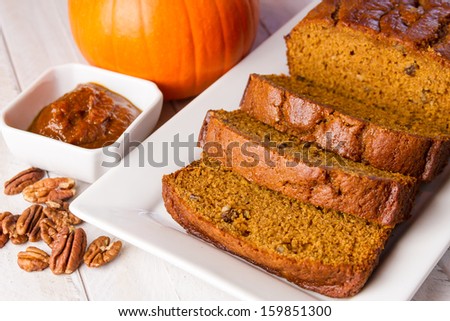 Home baked loaf of sliced pumpkin bread sitting on white plate with container of pumpkin butter and pecan nuts