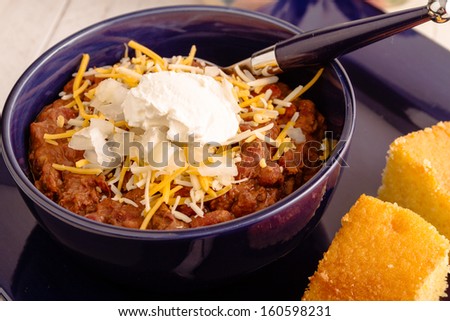 Close up of meat chili with red kidney beans, shredded cheese and sour cream in blue bowl with cornbread