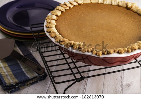 Fresh baked pumpkin pie sitting on white wooden table on wire baking rack and stack of colorful plates