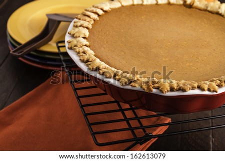 Fresh baked pumpkin pie sitting on wooden table on wire baking rack and stack of colorful plates