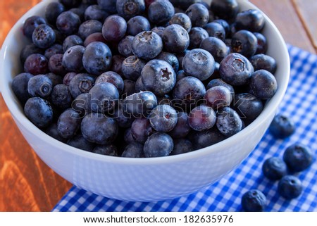 Close up of white bowl full of fresh organic blueberries on wooden table with blue gingham napkin