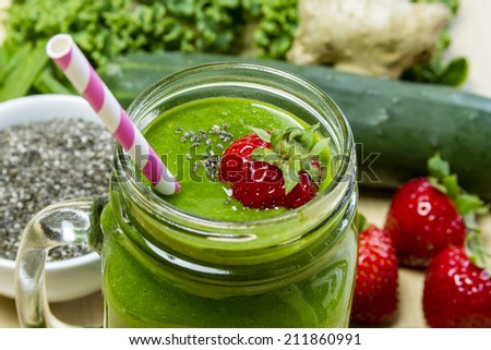 Healthy green juice smoothie surrounded by whole fruits, vegetables and chia seeds with fresh strawberry garnish and pink swirl straw