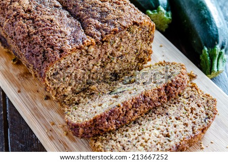 Loaf of homemade zucchini bread sitting on wooden cutting board with fresh zucchini squash
