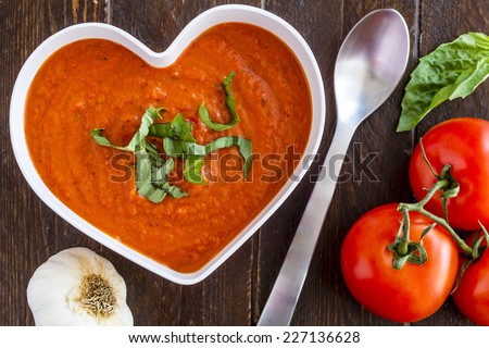 Homemade tomato and basil soup in white heart shaped bowl with spoon surrounded by fresh vegetables