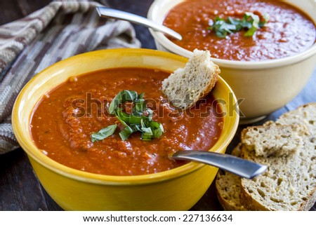 2 bowls of homemade tomato basil soup with spoons sitting on wooden table with brown striped napkin and slices of whole grain bread