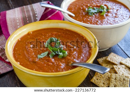 2 bowls of homemade tomato basil soup with spoons sitting on wooden table with red striped napkin and whole wheat crackers