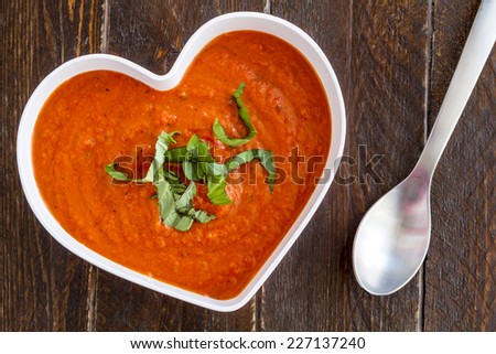 Homemade tomato and basil soup in white heart shaped bowl with spoon, whole wheat crackers, heirloom tomatoes and red heart napkin