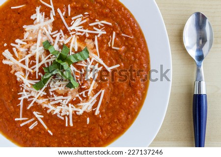 Homemade tomato and basil soup sprinkled with parmesan cheese in white round bowl with blue spoon