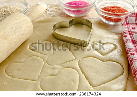Heart shaped cookie cutters cutting out holiday sugar cookies with wooden rolling pin and red and pink sugar sprinkles sitting next to red heart napkin