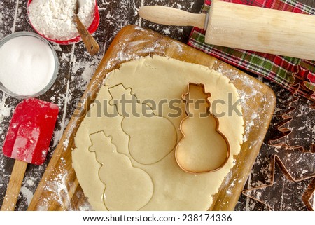 Copper cookie cutters cutting out holiday sugar cookies with wooden rolling pin and red spatula sitting on dark wood table