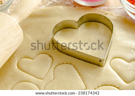 Close up of heart shaped cookie cutters cutting out holiday sugar cookies with wooden rolling pin and red and pink sugar sprinkles