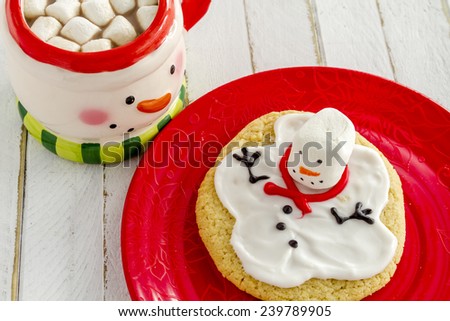 Melting snowman sugar cookie sitting on red plate with snowman mug filled with hot chocolate and marshmallows