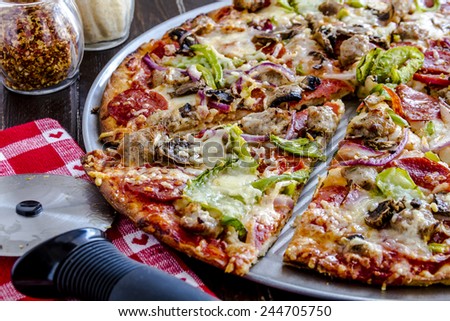 Close up of fresh baked thin crust supreme pizza surrounded by containers of parmesan cheese, red pepper flakes with red heart napkin and pizza cutter