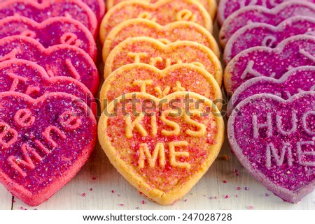 Rows of conversation heart cookies in pink, orange and purple with various text messages and sugar sprinkles