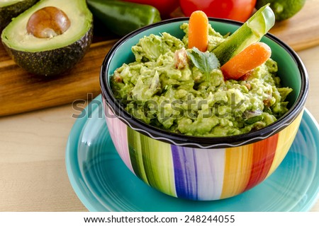 Fresh chunky guacamole in colorful bowl sitting on bright blue plate garnished with raw carrots and green peppers and cilantro