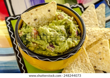 Close up of homemade chunky guacamole in bright yellow bowl sitting on colorful plate with white corn tortilla chips