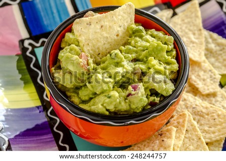 Close up of homemade chunky guacamole in bright orange bowl sitting on colorful plate with white corn tortilla chips