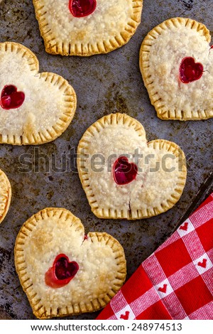 Heart shaped cherry hand pies lined up on metal baking pan with red and white heart kitchen towel