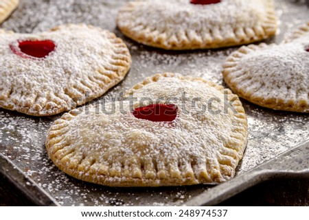Close up of heart shaped cherry hand pies dusted with powdered sugar sitting on metal baking pan