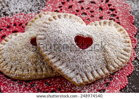 Close up of two heart shaped cherry hand pies dusted with confectioners sugar sitting on red lace doily