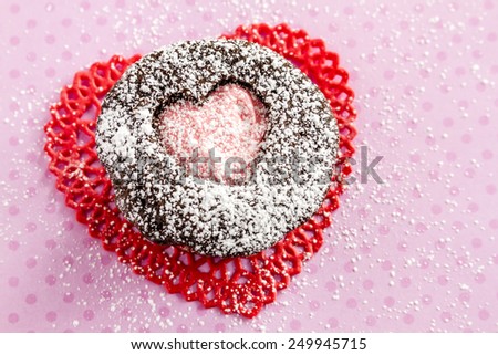 Chocolate cupcake with heart shaped cutout on top filled with pink frosting sitting on red lace doily on pink polka background dusted with powdered sugar