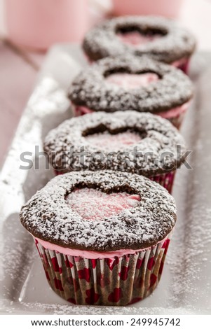 Line of chocolate cupcakes with heart shaped cutout filled with pink frosting sitting on white plate dusted with powdered sugar