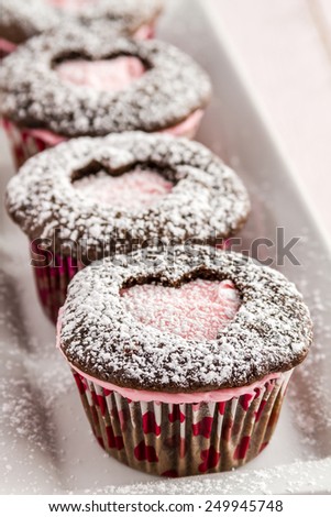 Close up of line of chocolate cupcakes with heart shaped cutout filled with pink frosting sitting on white plate dusted with powdered sugar