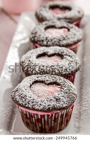 Line of chocolate cupcakes with heart shaped cutout filled with pink frosting sitting on white plate dusted with powdered sugar