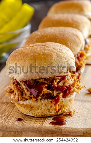 Close up of row of pulled pork barbeque sliders sitting on wooden cutting board with red striped napkin and dill pickles