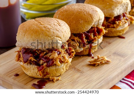 Row of pulled pork barbeque sliders sitting on wooden cutting board with red striped napkin and dill pickles in background