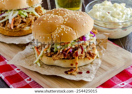 Two pulled pork barbeque sandwiches with coleslaw sitting on wooden cutting board with beer and potato salad