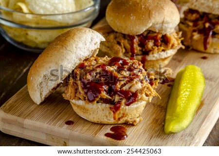 Three pulled pork barbeque sliders sitting on wooden cutting board with dill pickle and bowl of potato chips