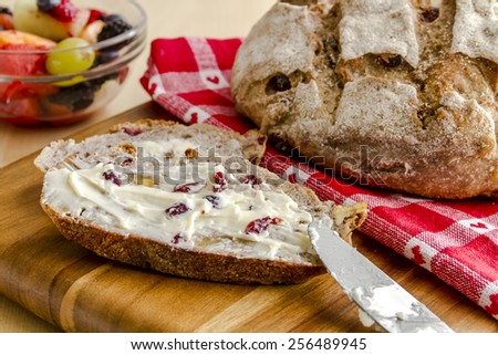 Whole loaf of walnut cranberry bread sitting on red heart napkin on wooden cutting board with slices of bread, butter, knife and fruit in bowl